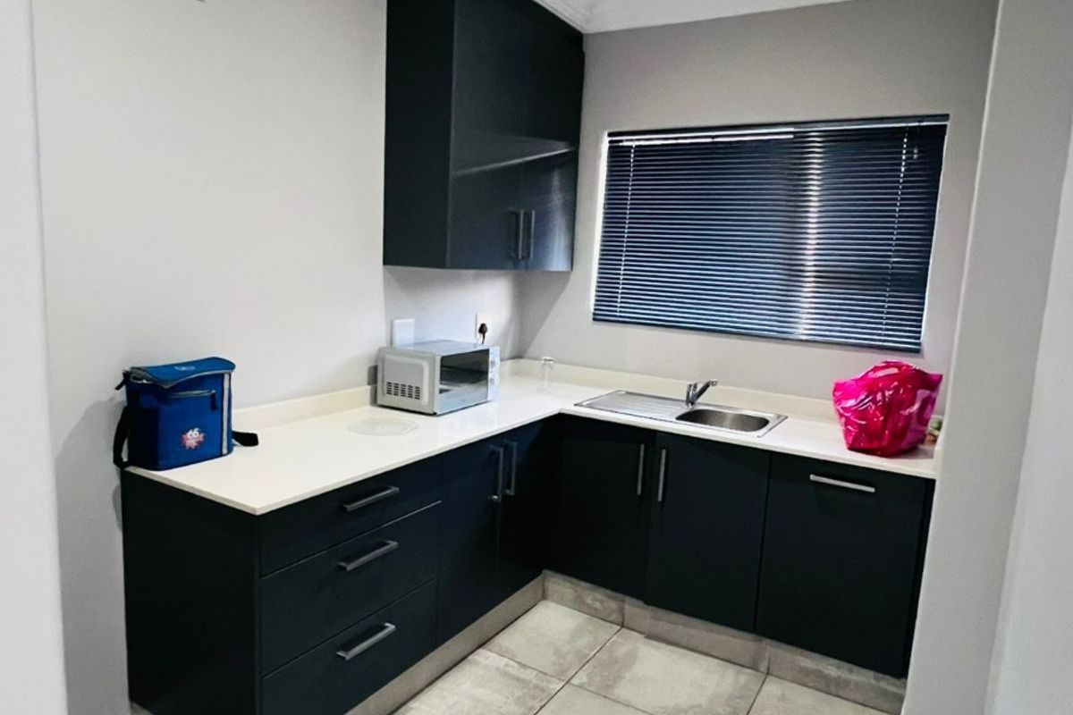 A modern, compact kitchenette featuring dark cabinetry, stone countertops, a stainless steel sink, and neutral floor tiling.