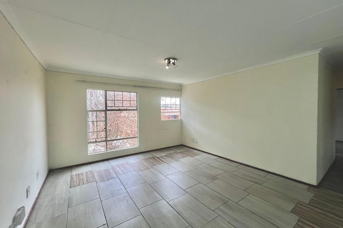 A spacious, empty living room featuring neutral tiled flooring, large windows providing natural light, and a simple ceiling-mounted light fixture.