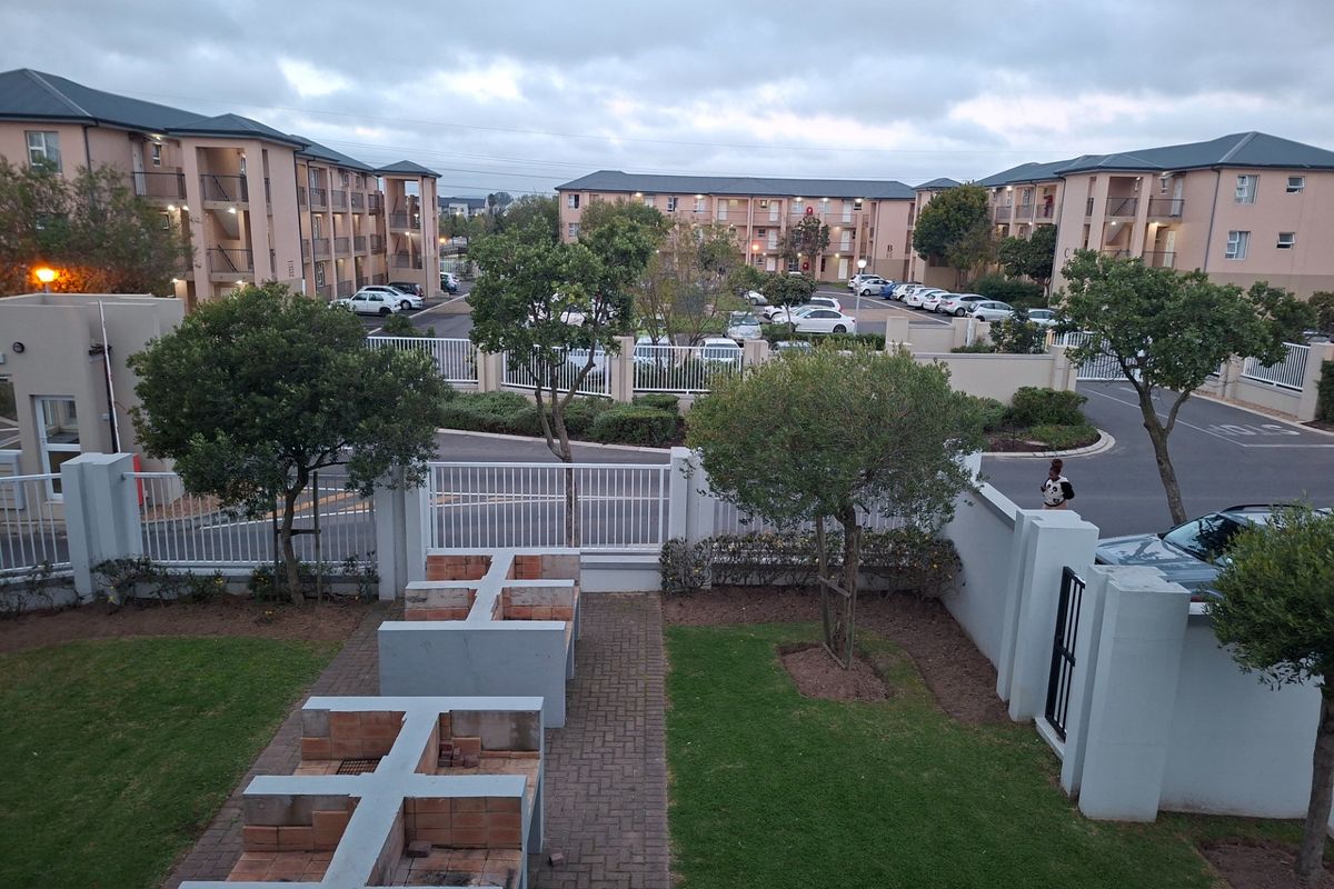A communal outdoor area featuring built-in brick braai facilities, paved pathways, and manicured lawns within a secure apartment complex setting.