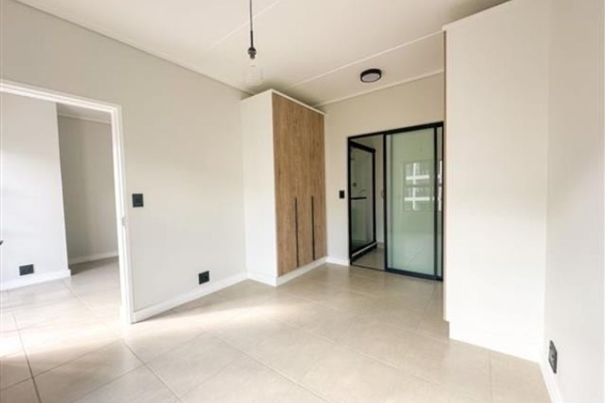 A bright, modern bedroom featuring neutral tiled flooring, built-in wood-finish cabinetry, and a sliding glass door leading to an en-suite bathroom.