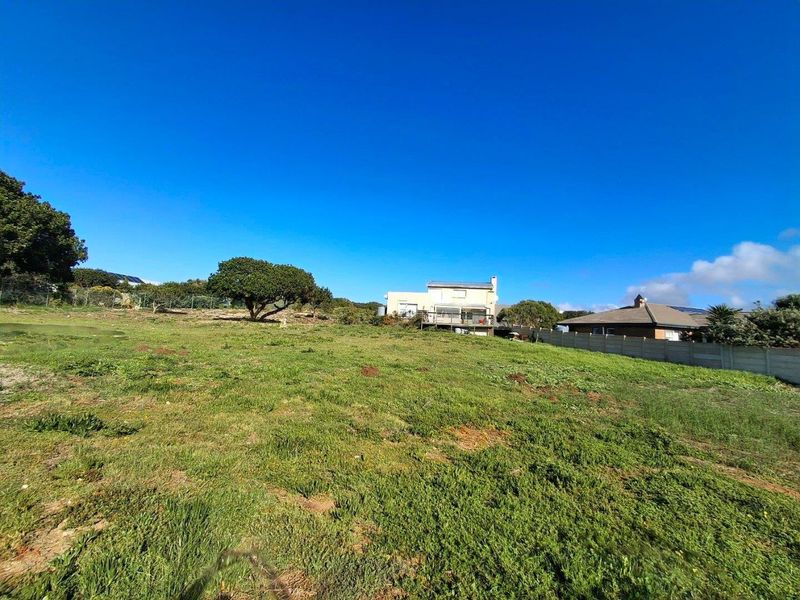 A wide-angle view of a vacant residential plot featuring natural grass coverage, perimeter fencing, and neighboring properties in the background.