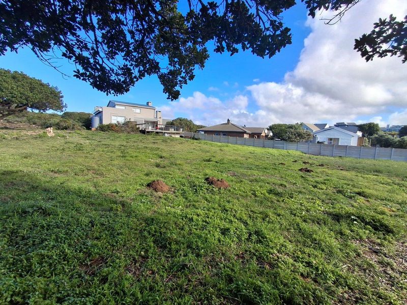 A view of the vacant residential land featuring grassy terrain, bordered by a perimeter fence with neighboring houses visible in the background.