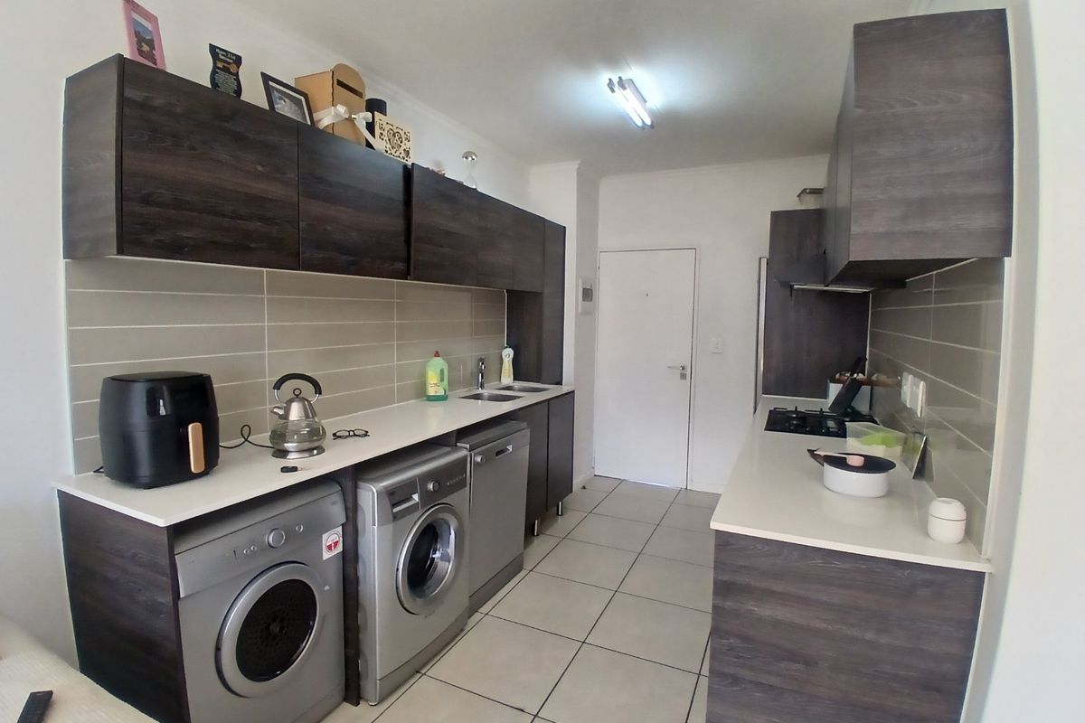 A functional galley-style kitchen featuring dark wood-grain cabinetry, light stone countertops, tiled backsplash, and integrated space for laundry appliances.