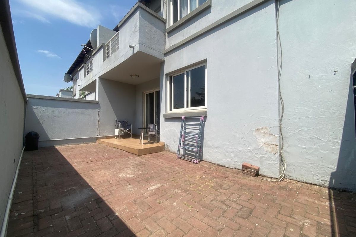 A rear exterior view of the townhouse featuring a paved courtyard, a small tiled patio area, and a balcony above.