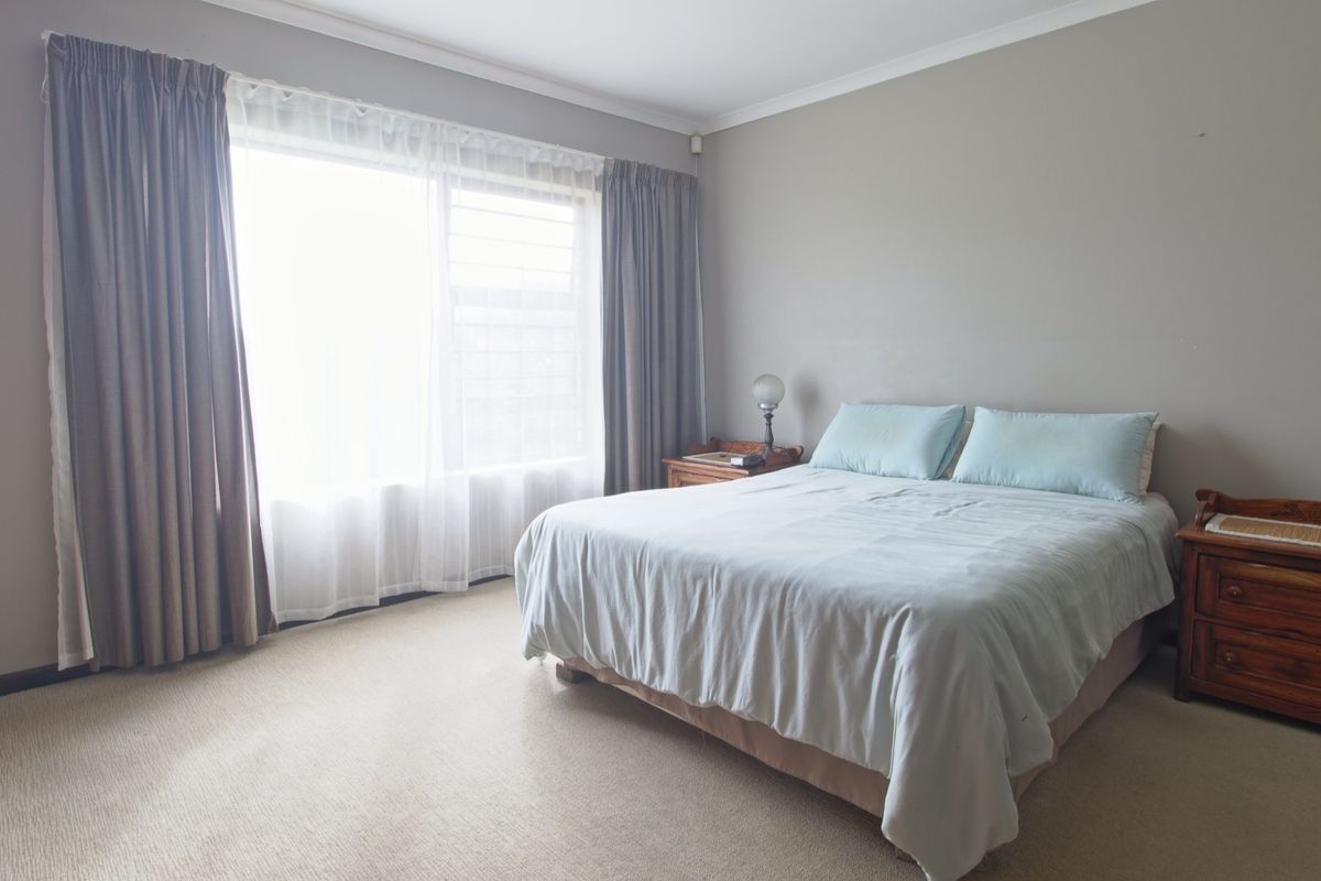 A well-lit bedroom featuring neutral wall tones, carpeted flooring, and a large window with curtain treatments.