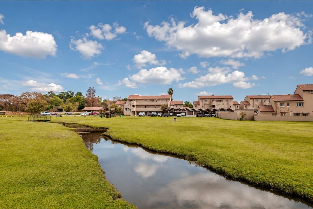 A wide-angle view of the apartment complex grounds, featuring a landscaped lawn, a small stream, and covered parking areas.