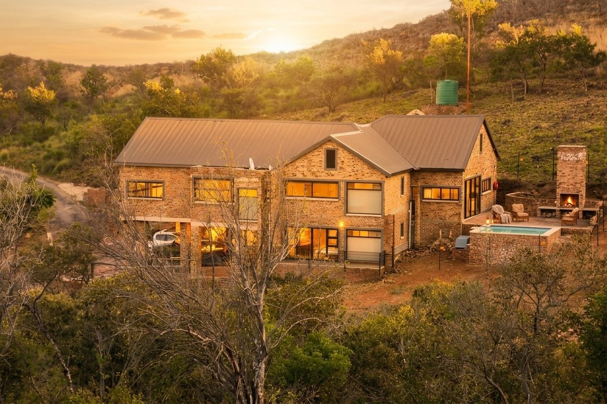 Aerial view of a multi-level brick house featuring a metal roof, swimming pool, and outdoor fireplace set in a natural landscape.