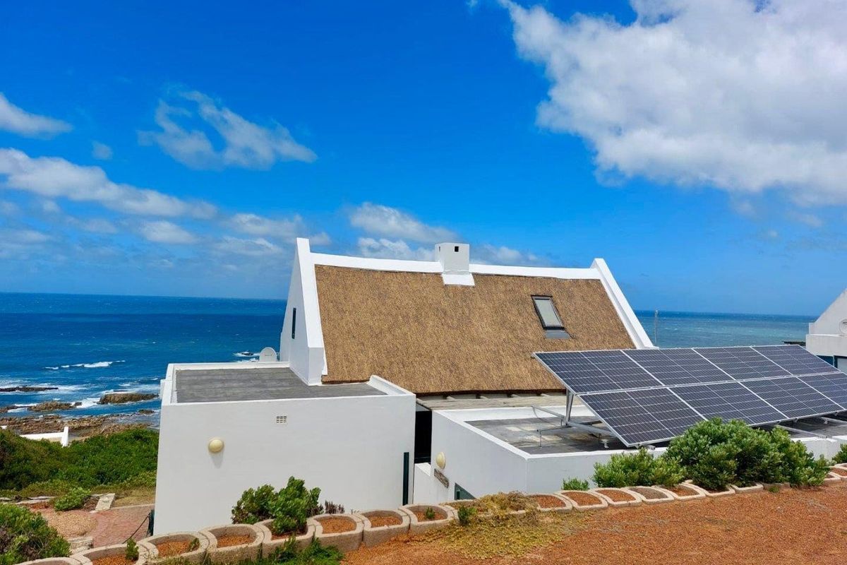 An elevated view of a coastal farm property featuring a traditional thatched roof, white exterior walls, and a modern solar panel installation.