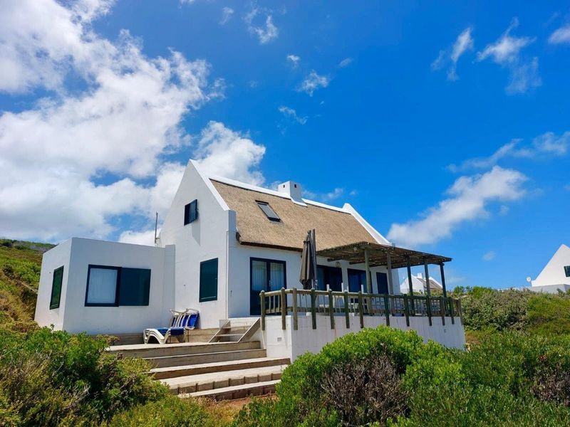 A charming white-walled farm cottage featuring a traditional thatched roof, a wooden deck, and a covered patio area set within a natural landscape.