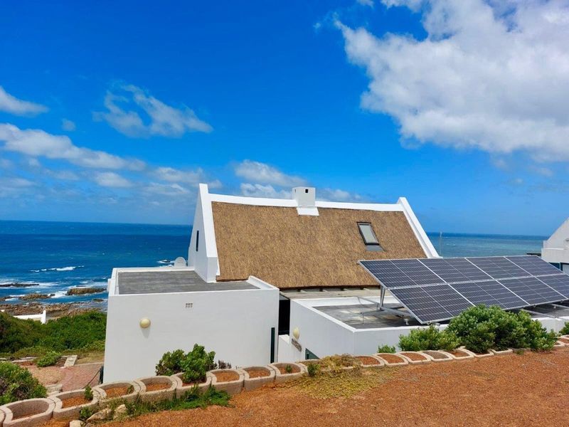 An elevated view of a coastal farm property featuring a traditional thatched roof, white exterior walls, and a modern solar panel installation.
