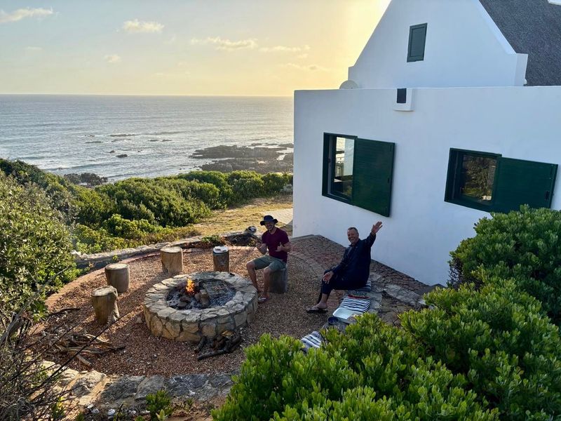 A scenic outdoor patio area featuring a stone-built fire pit, overlooking the ocean with direct access to the rear of the farmhouse.