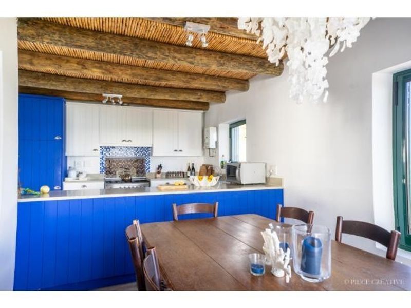 Open-plan kitchen and dining area featuring a blue cabinetry island, exposed timber beam ceiling, and tiled backsplash.