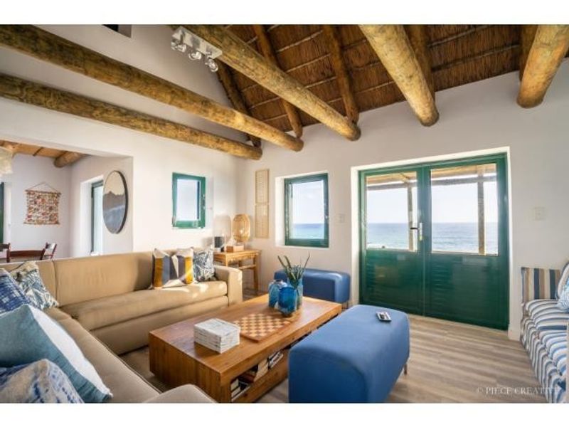 Bright living area featuring exposed timber rafters, a thatched ceiling, and double doors opening to a scenic coastal view.