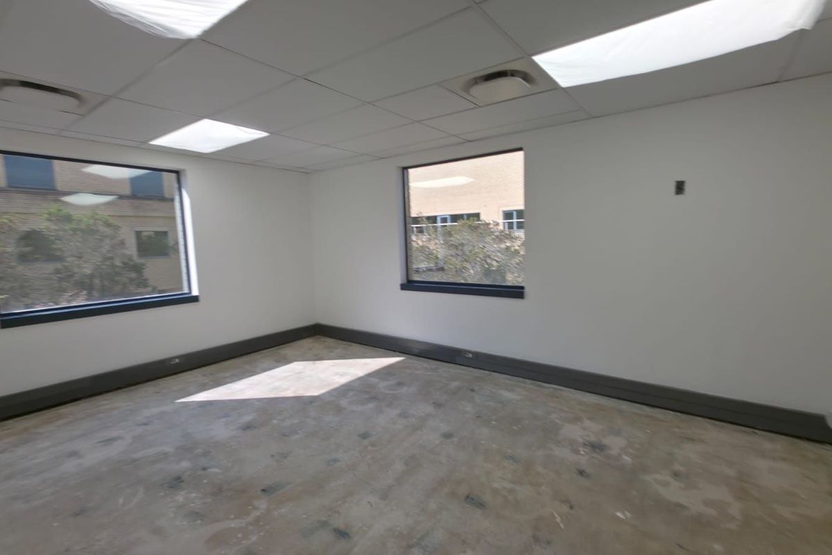An empty commercial office space featuring a suspended ceiling with recessed lighting, large windows, and unfinished concrete flooring.