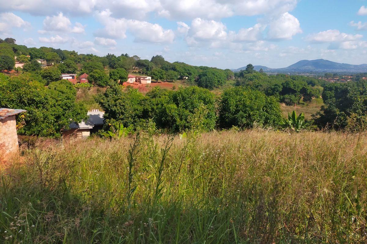 A scenic view overlooking a sloped, grassy residential plot with surrounding lush greenery and distant mountain vistas.