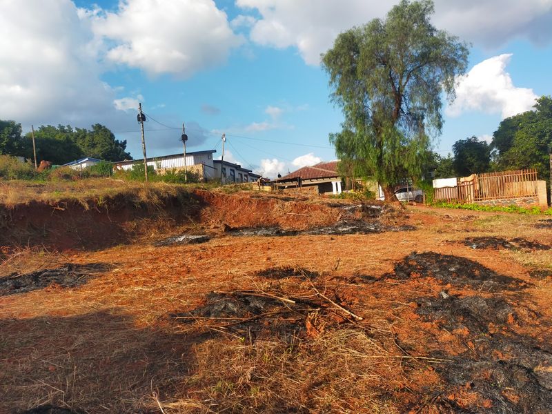 A vacant residential plot featuring uneven terrain, utility connections, and perimeter fencing, showing signs of recent vegetation clearing.
