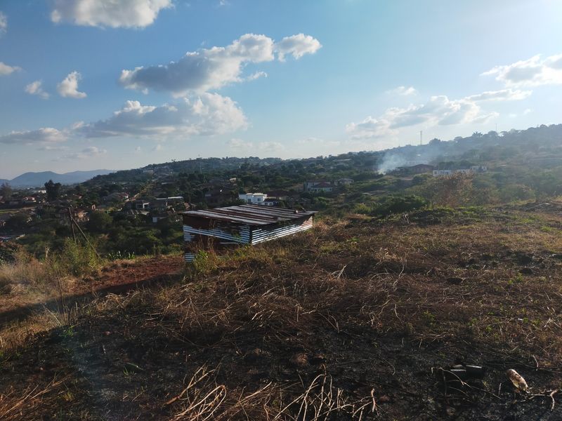 An elevated view of a vacant residential plot featuring a small corrugated metal shed and surrounding landscape with distant neighborhood views.