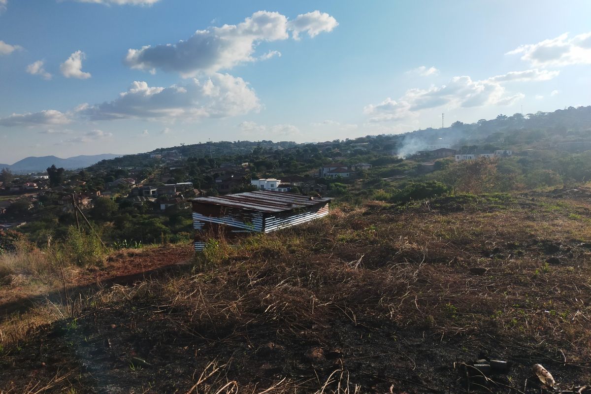 An elevated view of a vacant residential plot featuring a small corrugated metal shed and surrounding landscape with distant neighborhood views.