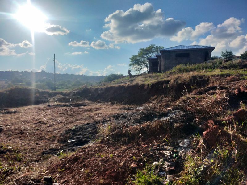 A view of a vacant residential plot featuring uneven terrain, scattered debris, and a utility pole with a distant structure visible.