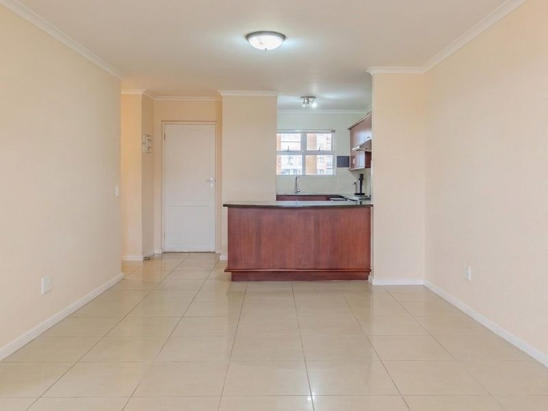An open-plan living and kitchen area featuring tiled flooring, a central kitchen island, and neutral wall finishes in a well-maintained apartment setting.