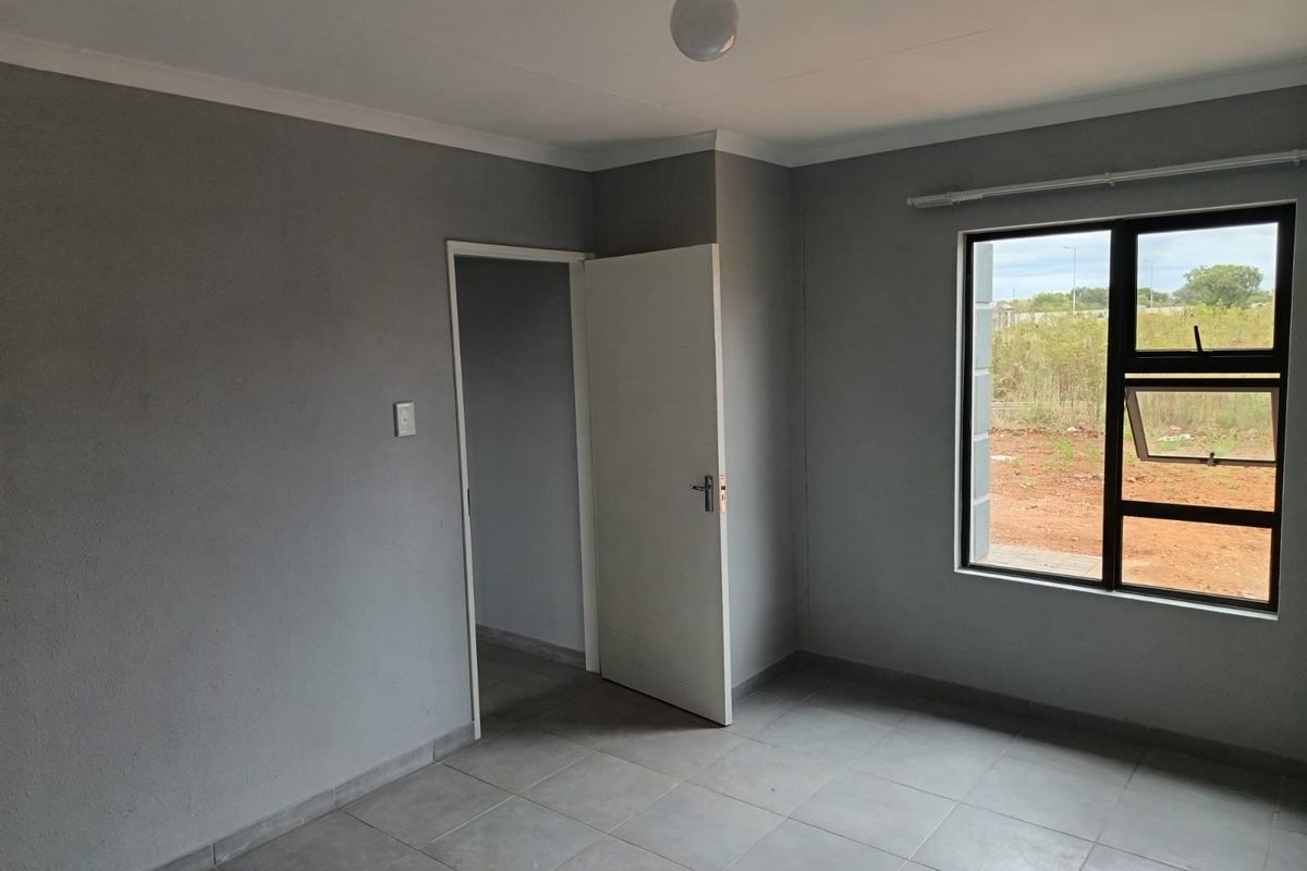 A clean, empty bedroom featuring neutral grey walls, tiled flooring, and a large window providing natural light and exterior views.
