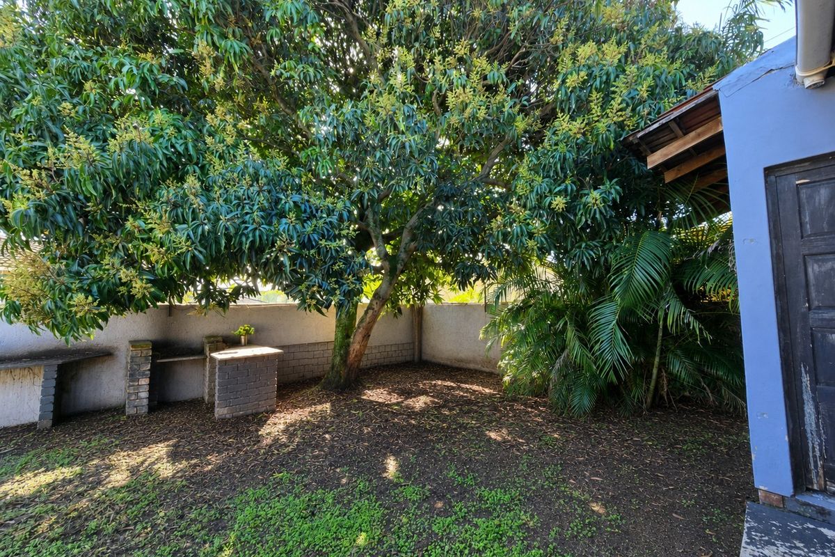 A private rear garden area featuring a built-in brick braai or barbecue station, mature trees, and a paved patio section.