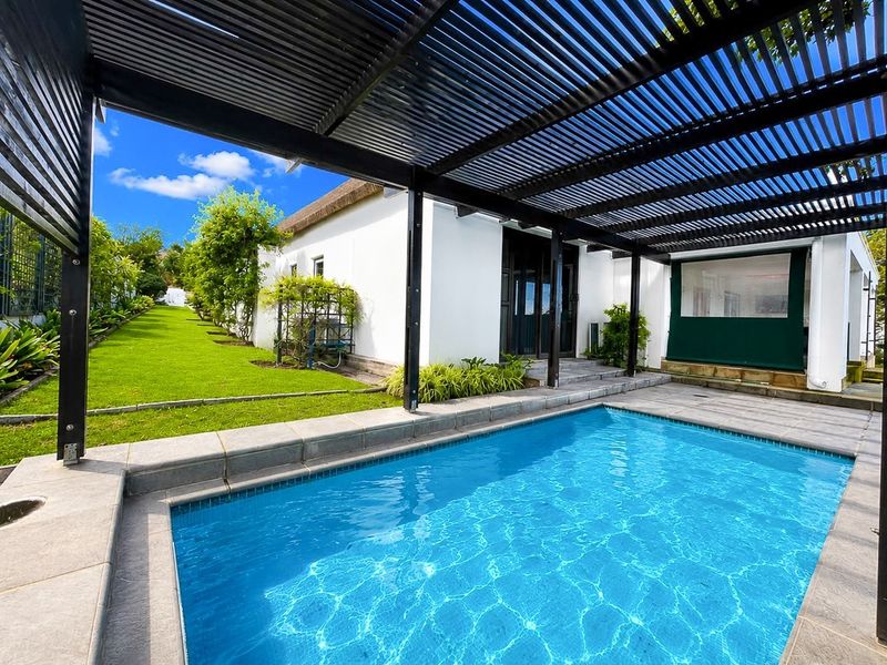 A well-maintained outdoor pool area featuring a modern pergola, paved decking, and a lush garden with a view of the main house.