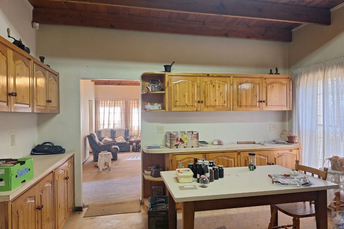 A dated kitchen featuring wooden cabinetry, exposed timber ceiling beams, and a laminate countertop, with an open-plan flow into the adjacent living area.