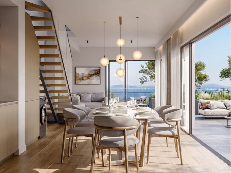 Modern open-plan living and dining area featuring timber flooring, a floating staircase, and large sliding glass doors opening to a scenic balcony with water views.