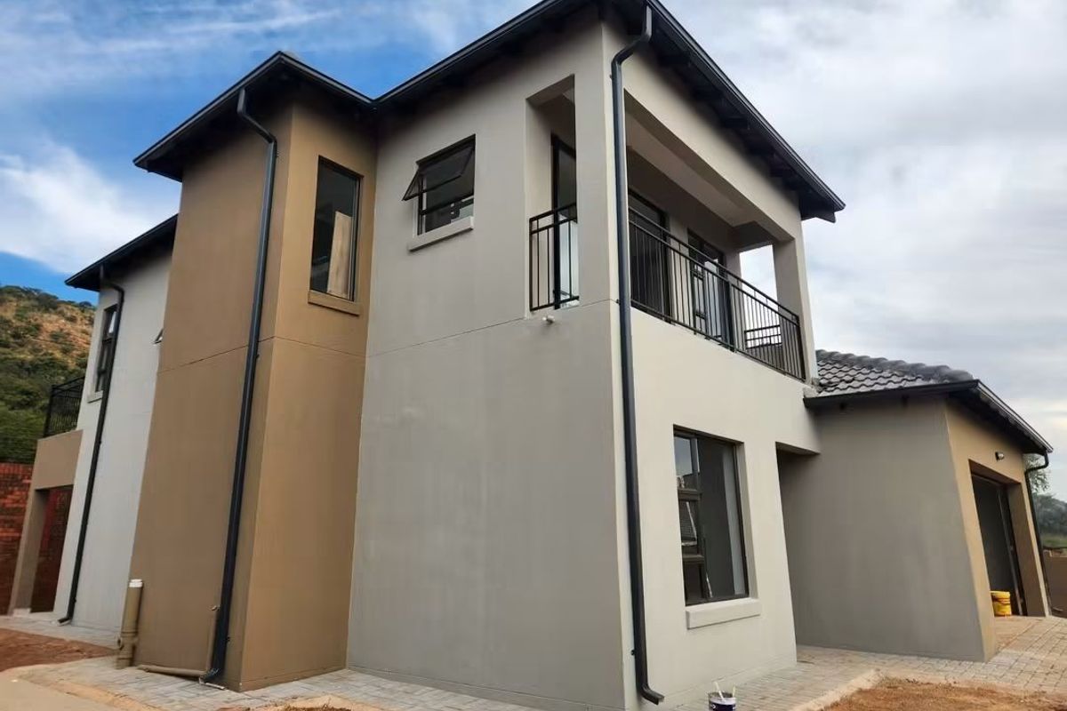 A modern two-story residential exterior featuring a neutral color palette, tiled roof, balcony with metal railing, and paved driveway area.