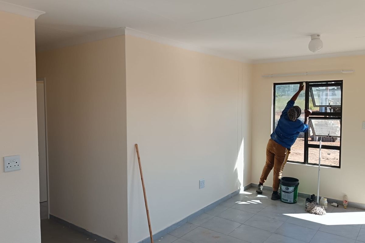 A bright, empty living area featuring neutral painted walls, tiled flooring, and modern aluminium window frames, currently undergoing final finishing touches.