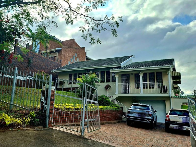 Front view of a multi-level residential property featuring a paved driveway, garage, elevated balcony, and landscaped garden with brick retaining walls.