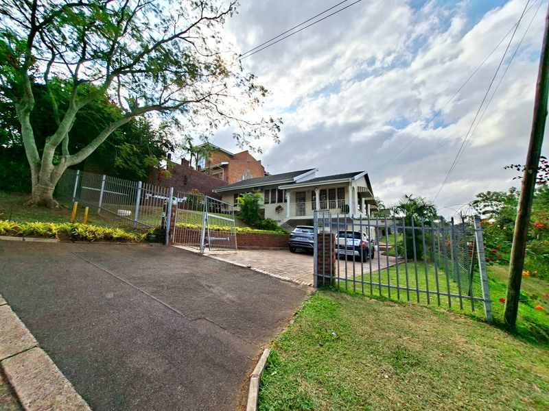 Front view of the property featuring a paved driveway, gated entrance, and established garden surroundings with a multi-level residential structure.