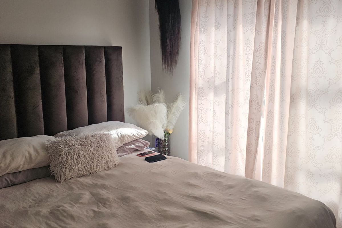 A bedroom featuring a dark upholstered headboard and natural light filtering through patterned curtains.