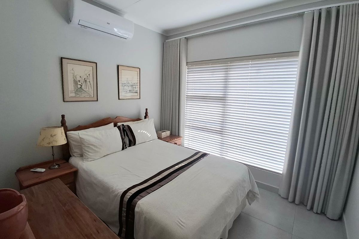 A well-maintained bedroom featuring tiled flooring, a wall-mounted air conditioning unit, and large windows with fitted blinds and curtains.