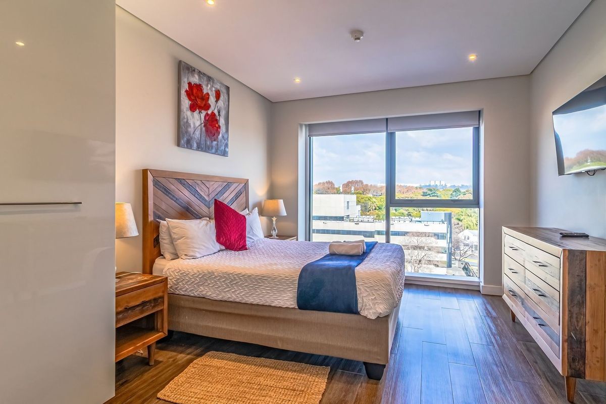 A modern, well-lit bedroom featuring large windows with city views, sleek built-in cabinetry, and high-quality wood-look flooring.