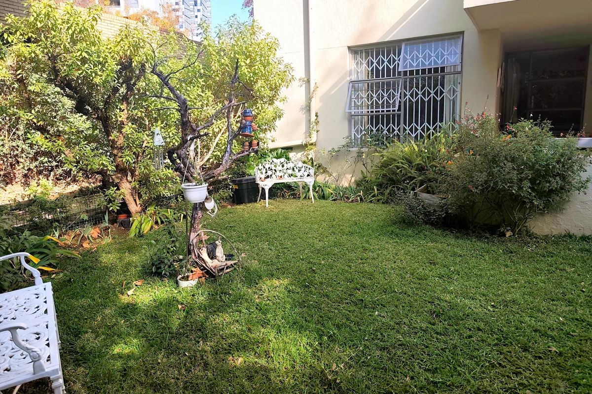 A private rear garden area featuring a lawn, mature trees, and direct access to the apartment building with security bars on the windows.