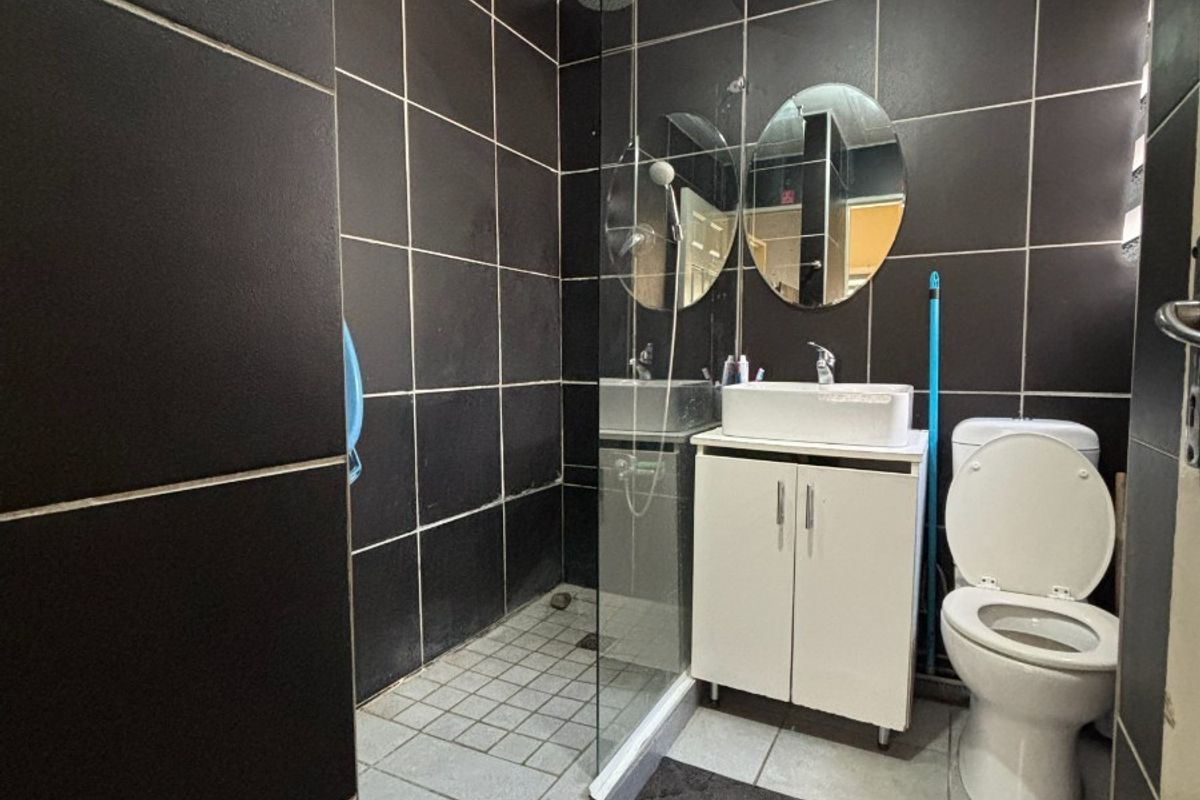 A compact bathroom featuring dark wall tiling, a glass-enclosed walk-in shower, a white vanity basin, and a standard toilet.