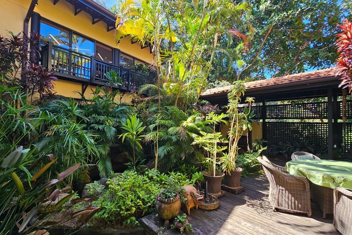 A lush, landscaped garden courtyard featuring a wooden deck, mature tropical plantings, and a covered patio area adjacent to the main house structure.