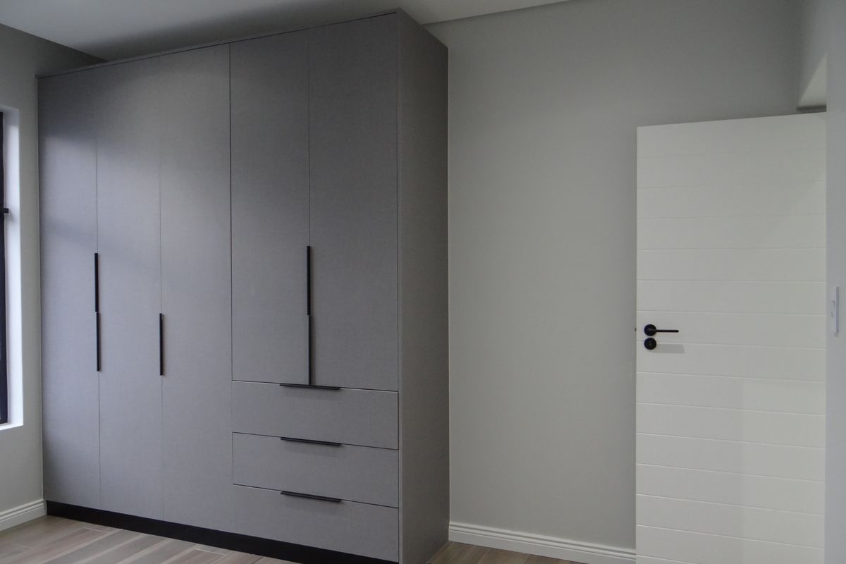 A modern bedroom featuring sleek, floor-to-ceiling grey built-in cabinetry with matte black hardware and light-toned wood-look flooring.