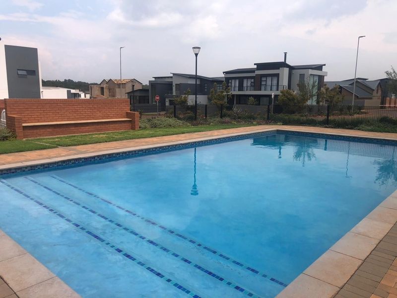 A well-maintained swimming pool area featuring paved surrounds, a brick feature wall, and landscaped garden elements in a residential estate setting.