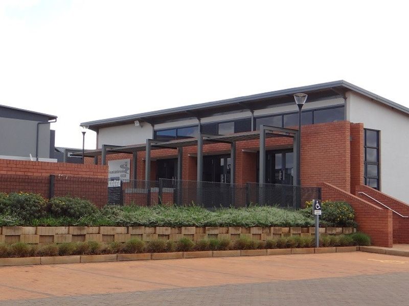 Exterior view of a modern brick clubhouse building featuring a covered entrance walkway, paved parking area, and landscaped retaining walls.