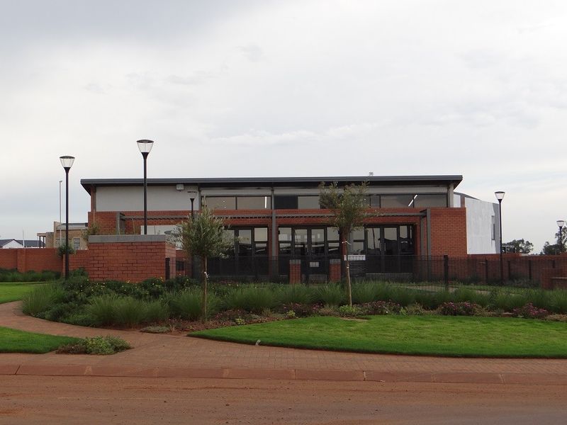 A modern clubhouse facility featuring face-brick exterior walls, large glass paneling, and a well-maintained landscaped garden area with paved pathways.