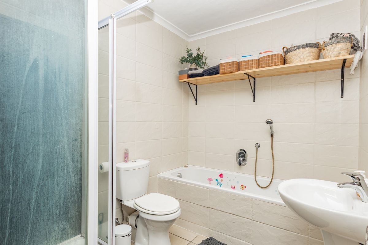 A functional bathroom featuring a combined shower and bathtub, pedestal basin, and toilet with neutral wall tiling and wall-mounted shelving.