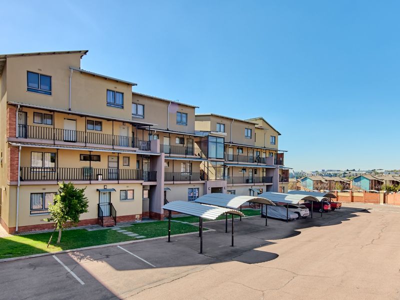 Exterior view of a multi-story apartment complex featuring private balconies, metal railings, and covered parking bays within a paved residential courtyard.