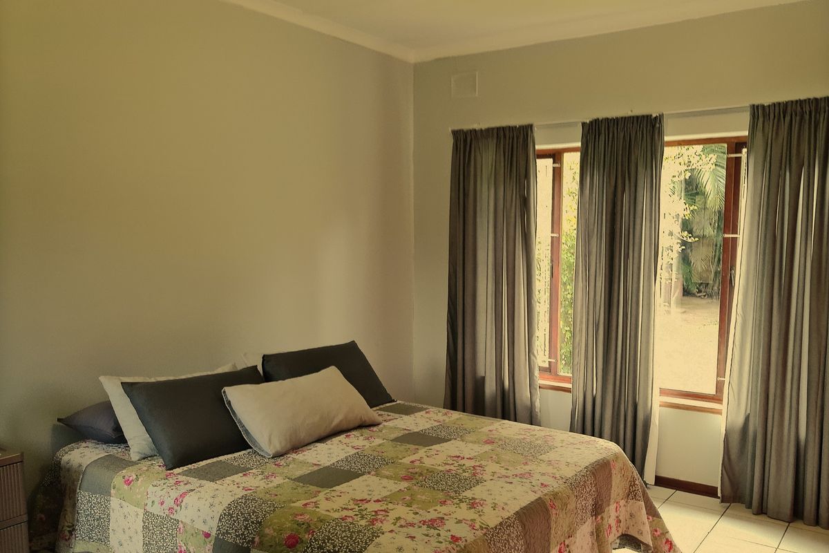 A bedroom featuring tiled flooring and large windows with curtains, providing natural light and a view of the garden area.