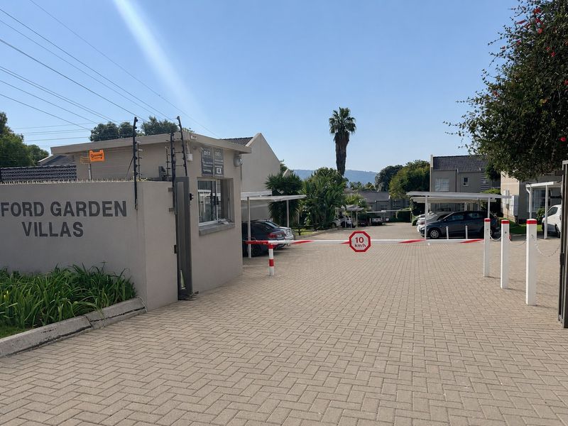 The entrance to Ford Garden Villas featuring a secure gated access point with a guardhouse, paved driveway, and covered parking bays.