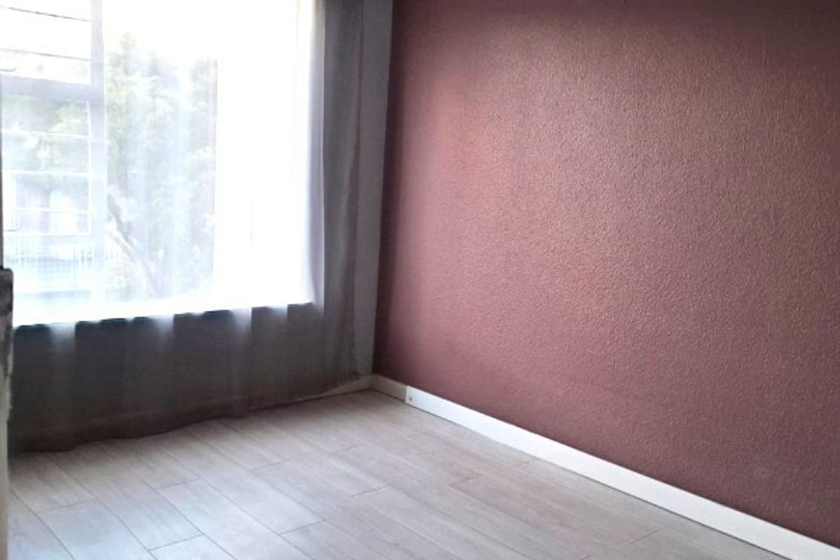 A bright, empty bedroom featuring light-toned wood-look flooring, a large window with sheer curtains, and a contrasting dark red accent wall.