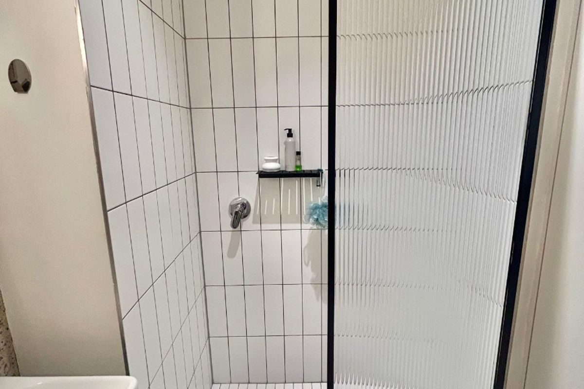 A modern bathroom shower featuring white vertical wall tiles, a grid-patterned floor, and a stylish black-framed fluted glass shower screen.