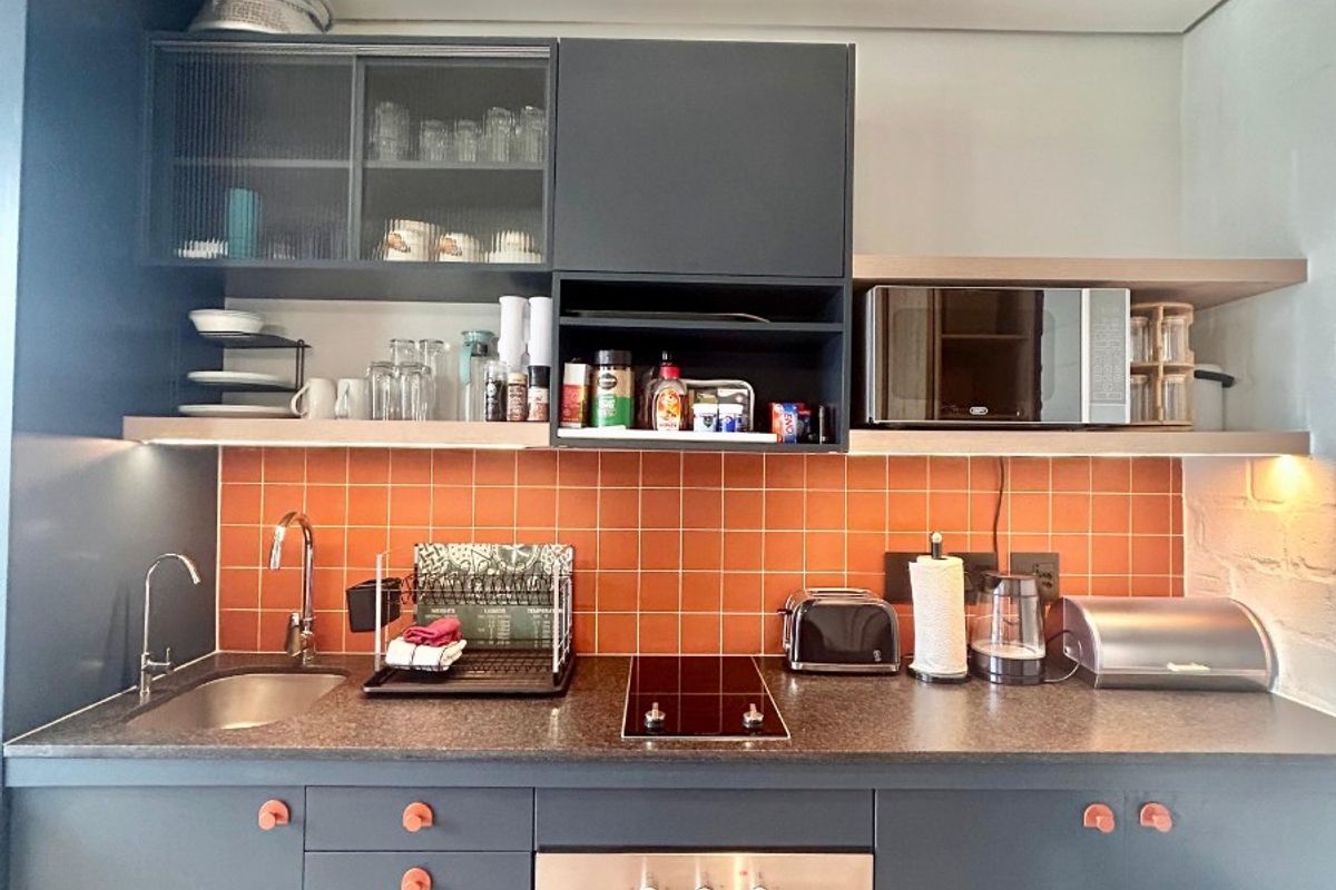 Modern kitchen featuring dark cabinetry, orange tiled backsplash, integrated oven, electric hob, and exposed concrete ceiling.